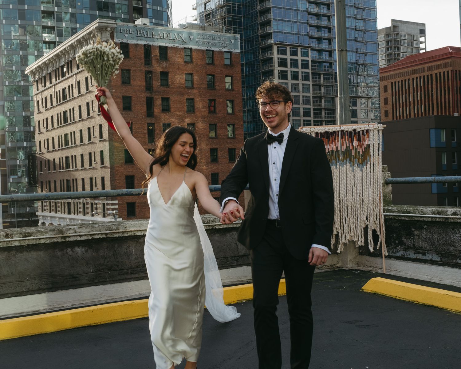 Joyful newlyweds celebrate on a rooftop in NYC, bride in white gown holding bouquet, groom in black tuxedo.
