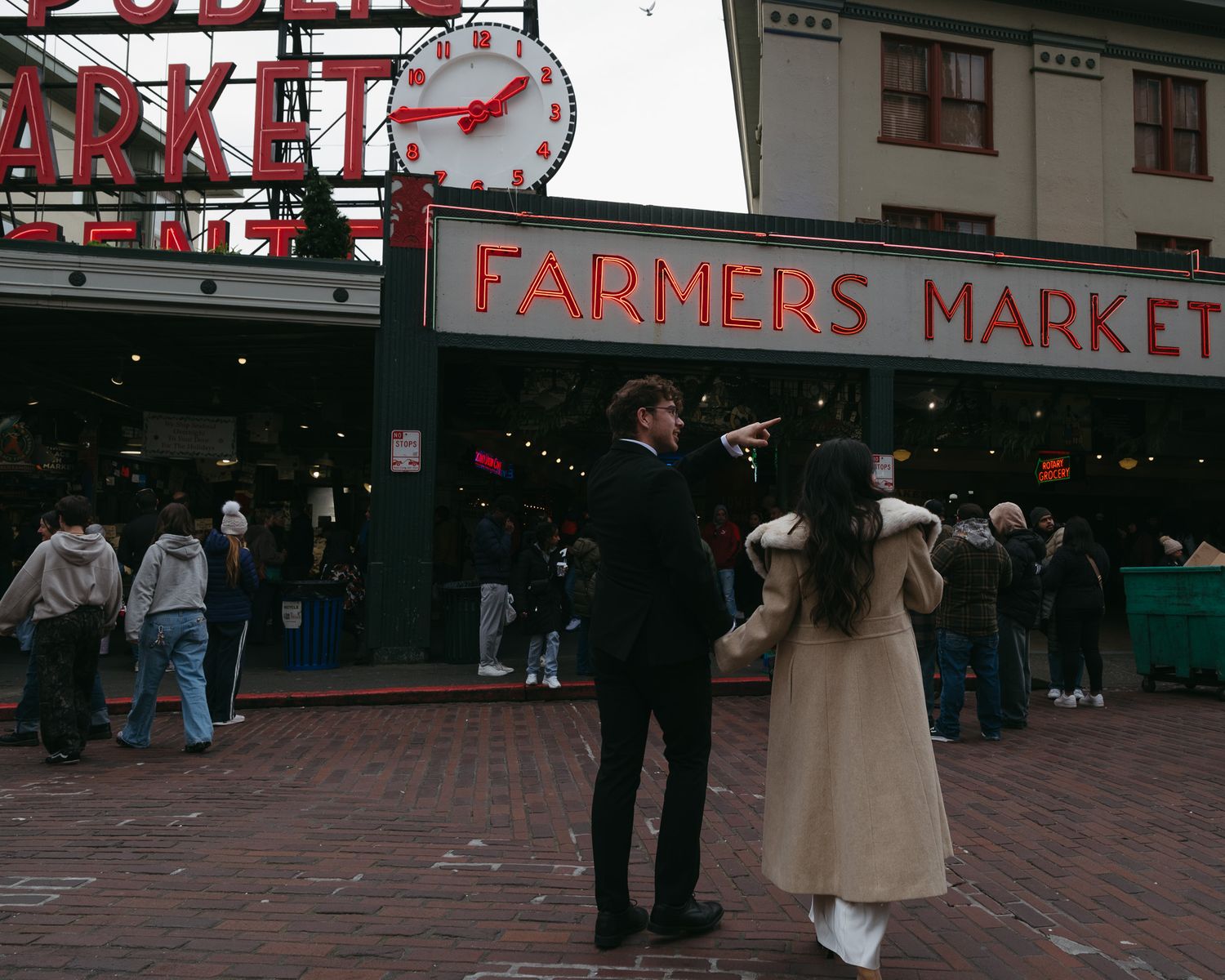 Couple in formal attire standing outside Pike Place Farmers Market in Seattle, with a brick plaza and crowd in the background.