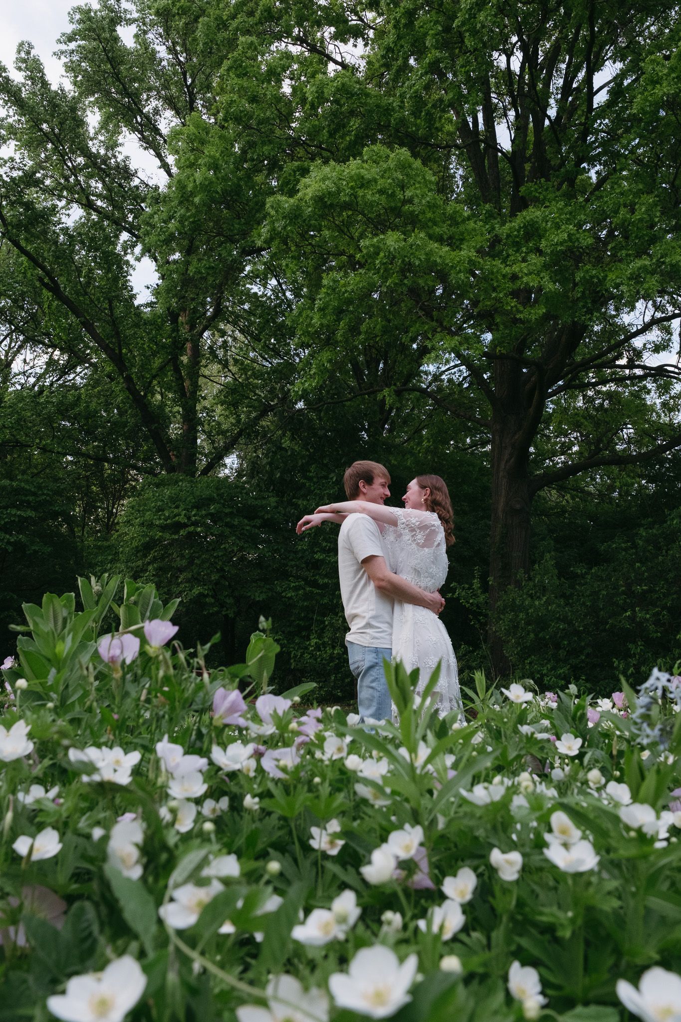 Couple embracing in a lush garden surrounded by white and pink flowers with tall green trees behind them.