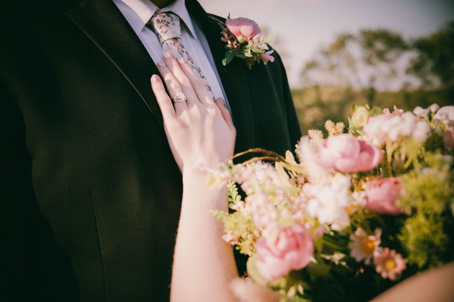 Bride holding pink peony bouquet with groom in black suit wearing floral tie and boutonniere at outdoor wedding.