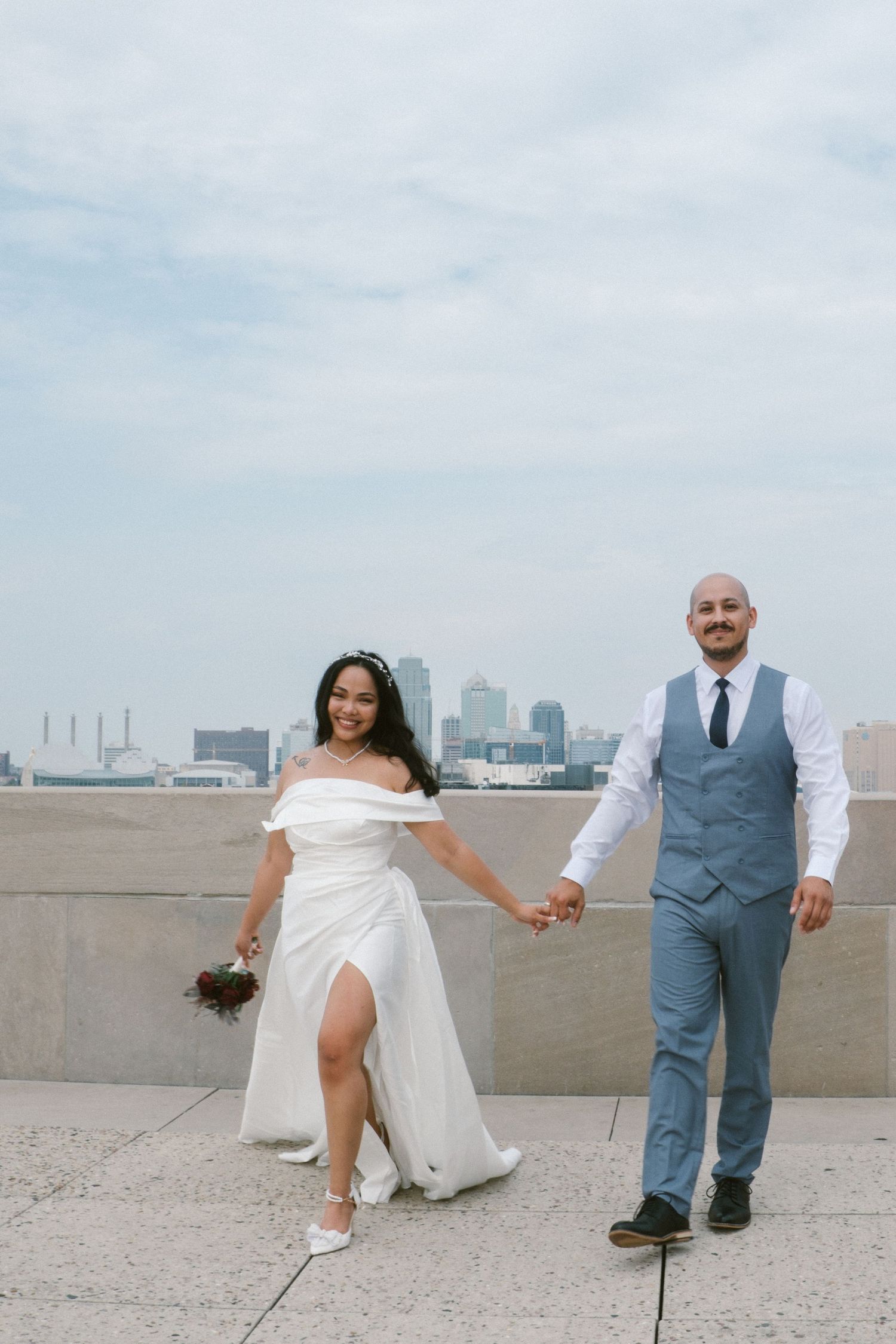 Bride and groom hold hands on a rooftop with city skyline backdrop, bride in off-shoulder white gown, groom in blue suit.