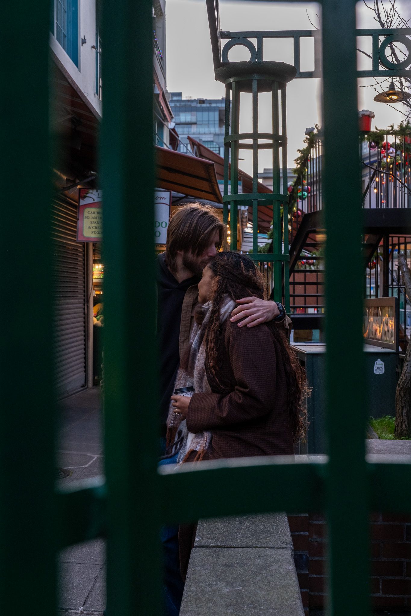 Couple embracing romantically on a city street, viewed through green metal fence bars at dusk.