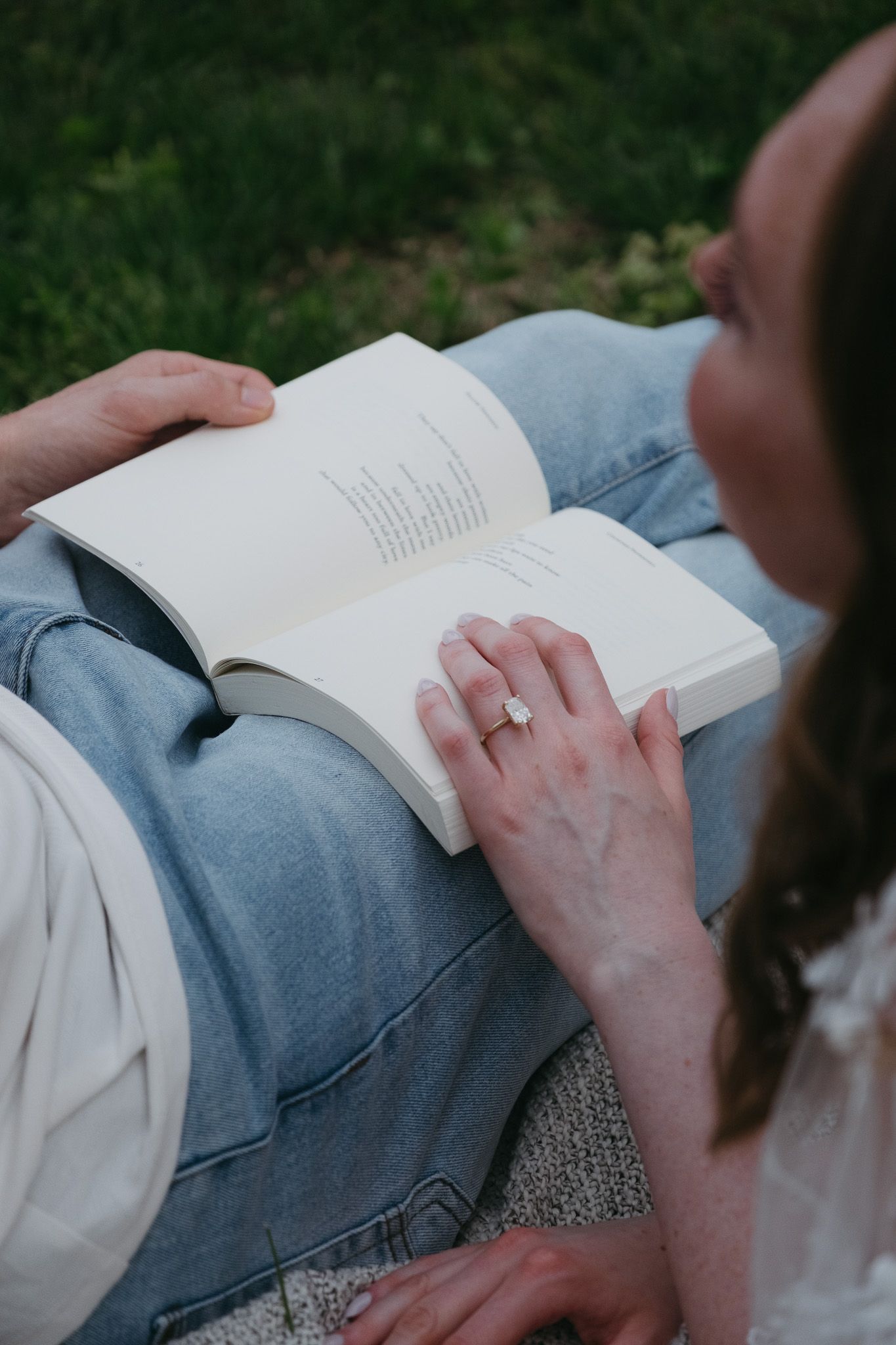 A woman with a ring reads a book outdoors on grass, leaning against someone in jeans on a sunny day.