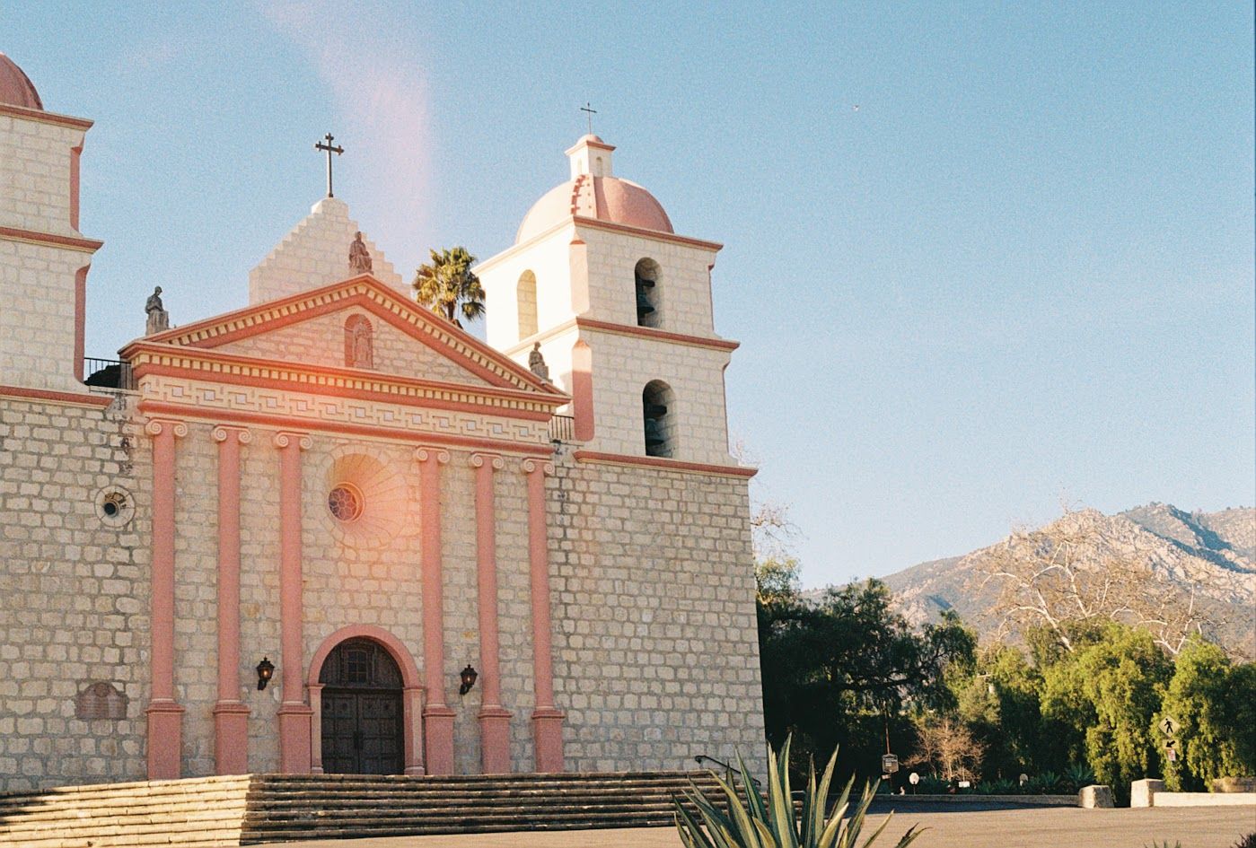 Historic Spanish colonial mission church with pink facade and white bell towers stands against blue sky and mountain backdrop.