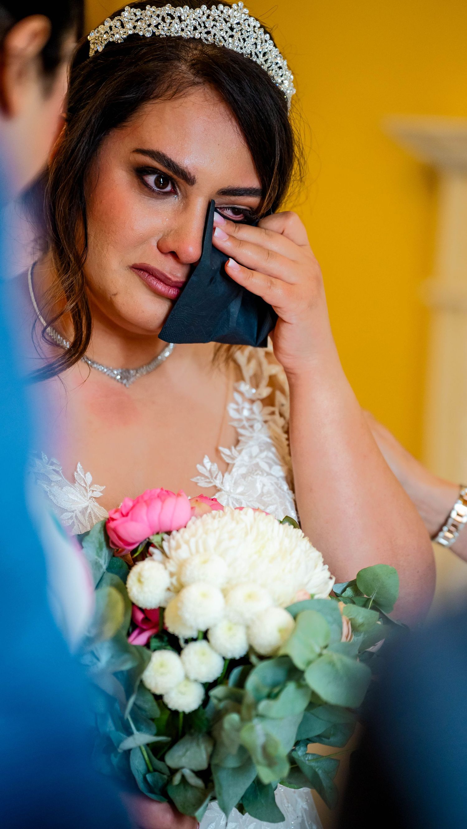 A bride wearing a sparkling tiara holds a bouquet of white roses against a golden background.