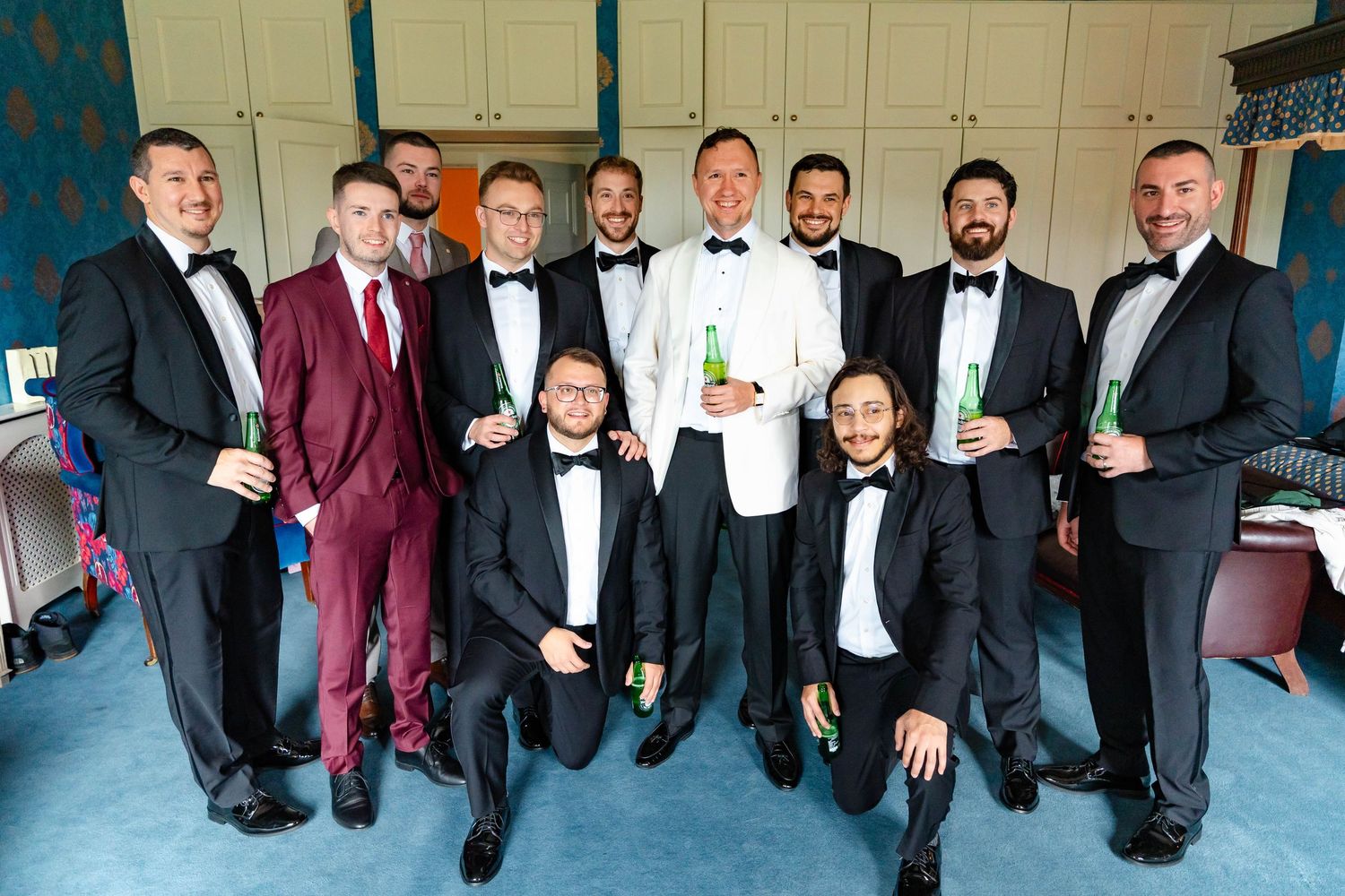 Groomsmen in black tuxedos and burgundy suits pose together for a formal wedding photo.