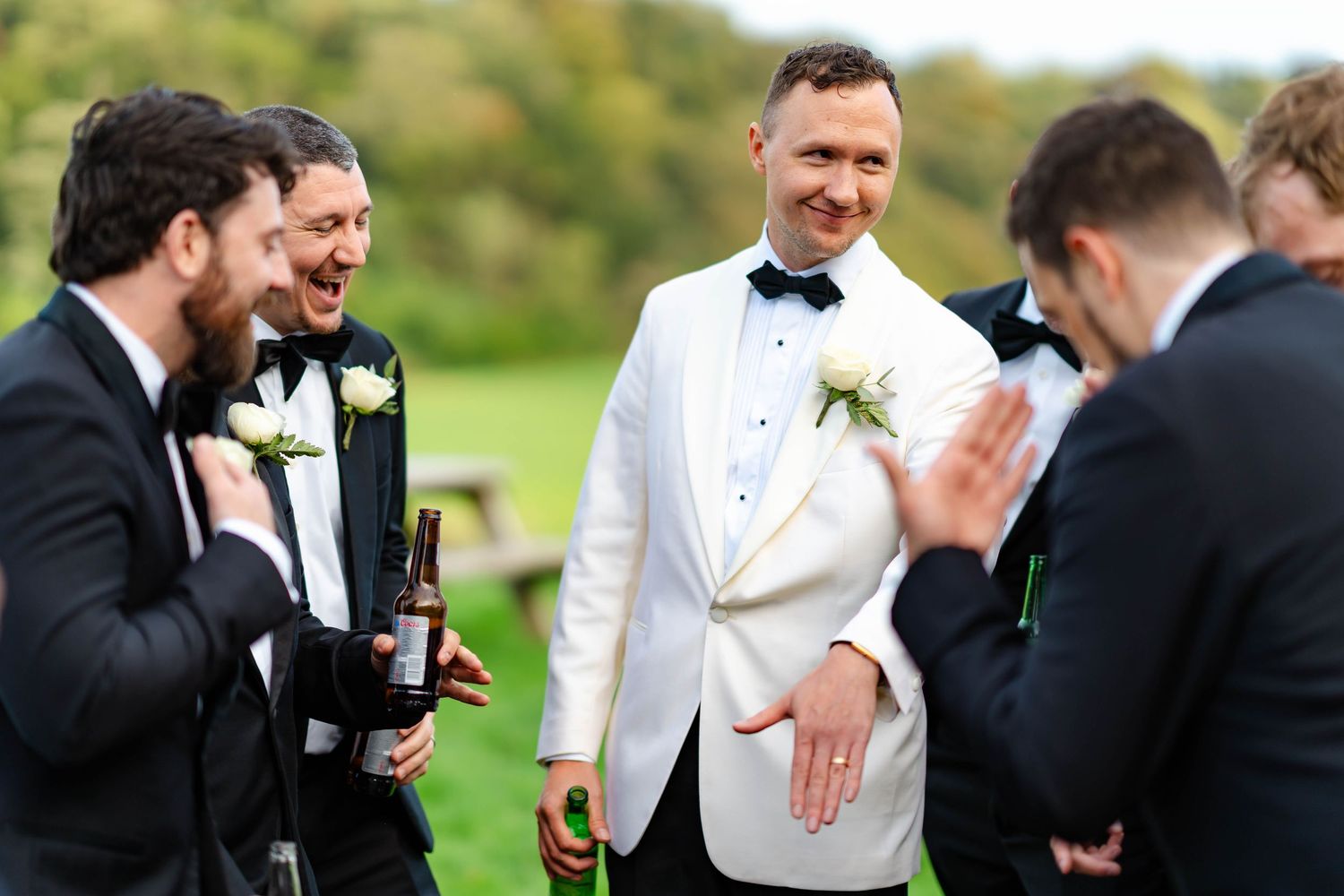Groomsmen in black and white tuxedos share joyful laughter during outdoor wedding photography session.