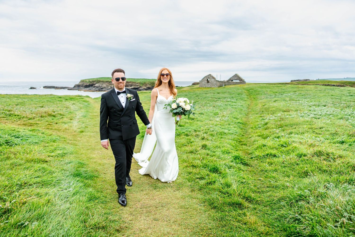 A couple strolls along a coastal path with green fields and ocean views.