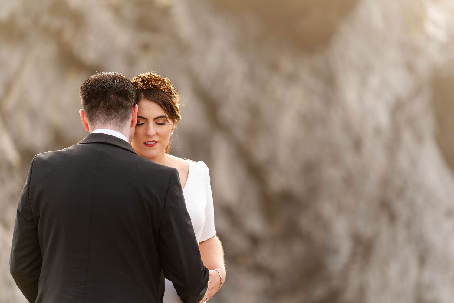 A couple embraces during a romantic wedding photoshoot against a textured stone wall.
