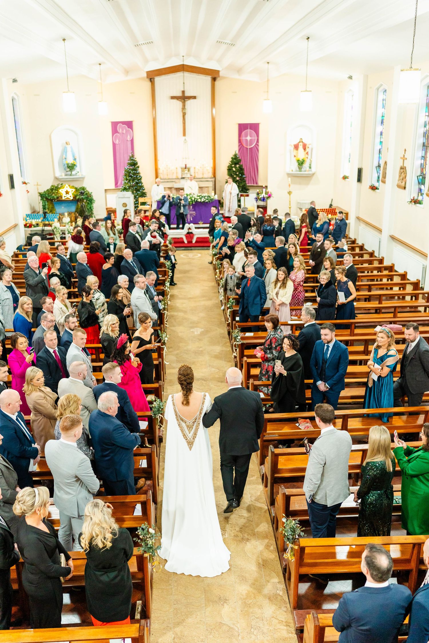Church wedding ceremony with guests standing in wooden pews and purple decorative elements.