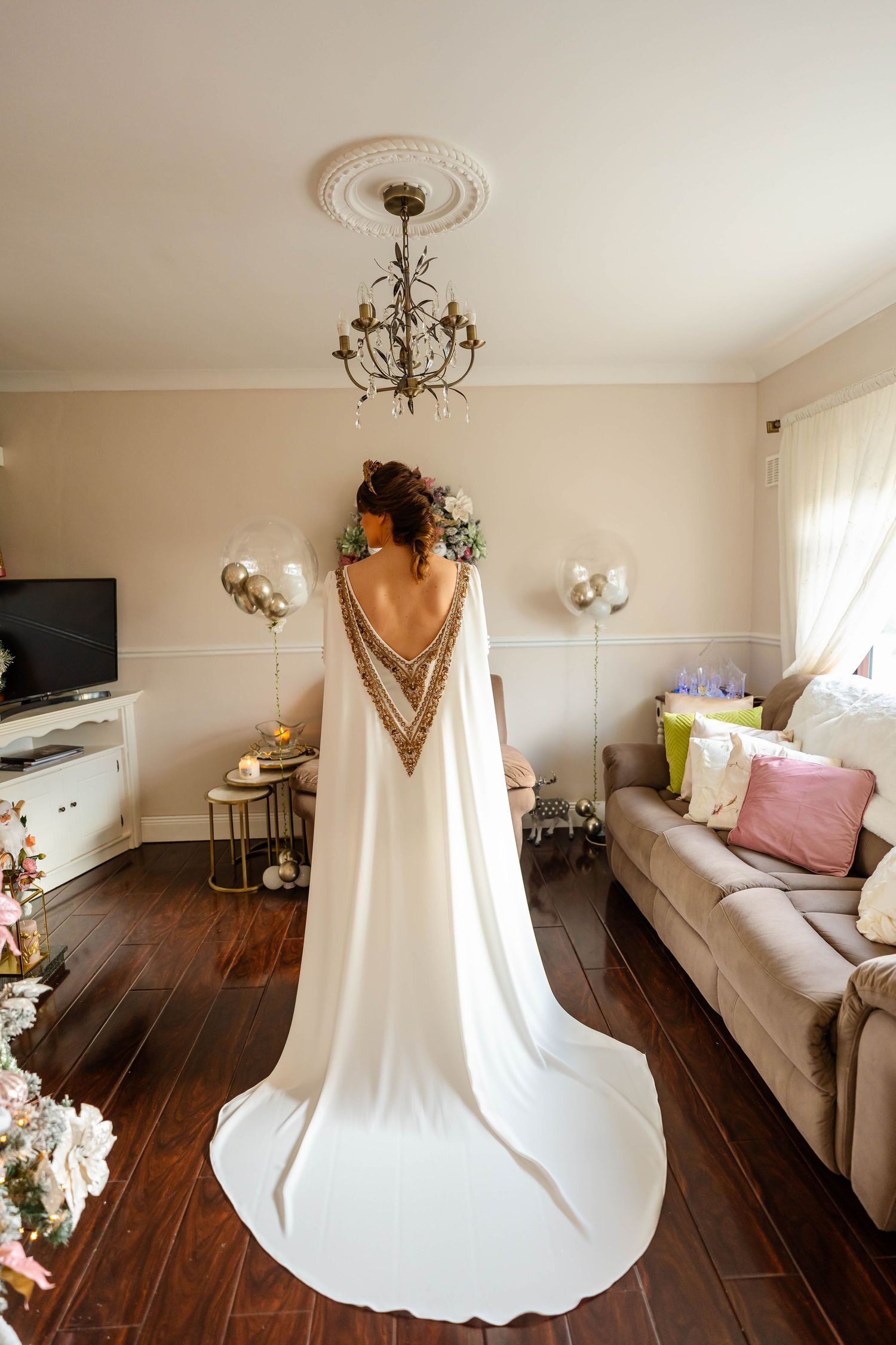 Elegant white wedding dress hanging in a bright room with a chandelier and dark wood floors.