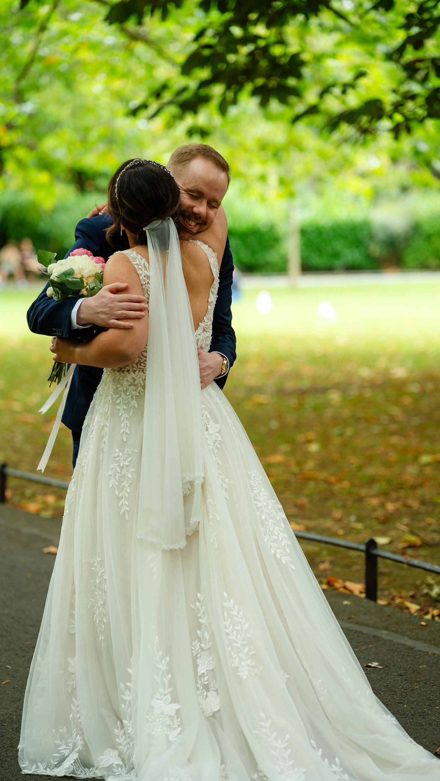 Couple embracing during their first look on a tree-lined path with fallen autumn leaves.