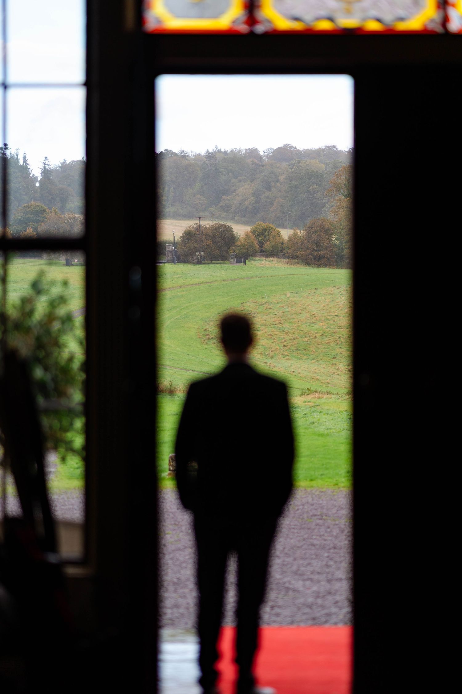 A silhouette stands in a doorway overlooking green countryside fields and distant trees on the horizon.