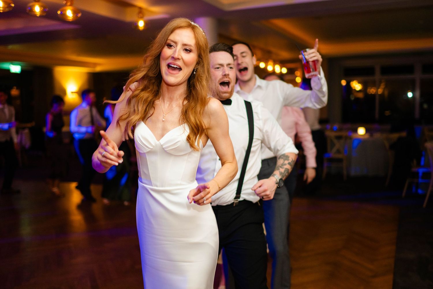Wedding celebration with guests dancing joyfully under warm reception lighting.