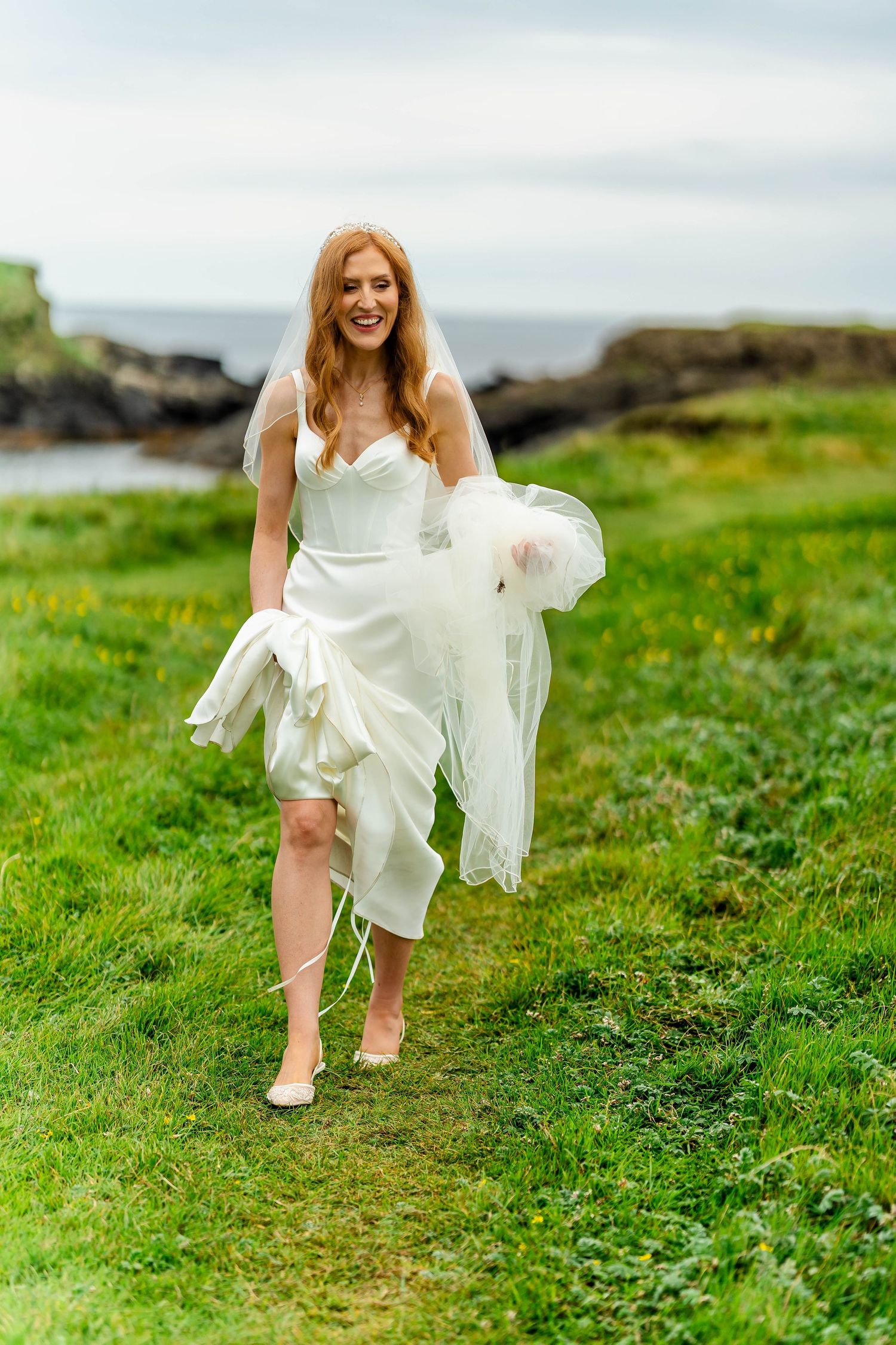 Playful bride lifting her white wedding dress while walking on coastal grass.