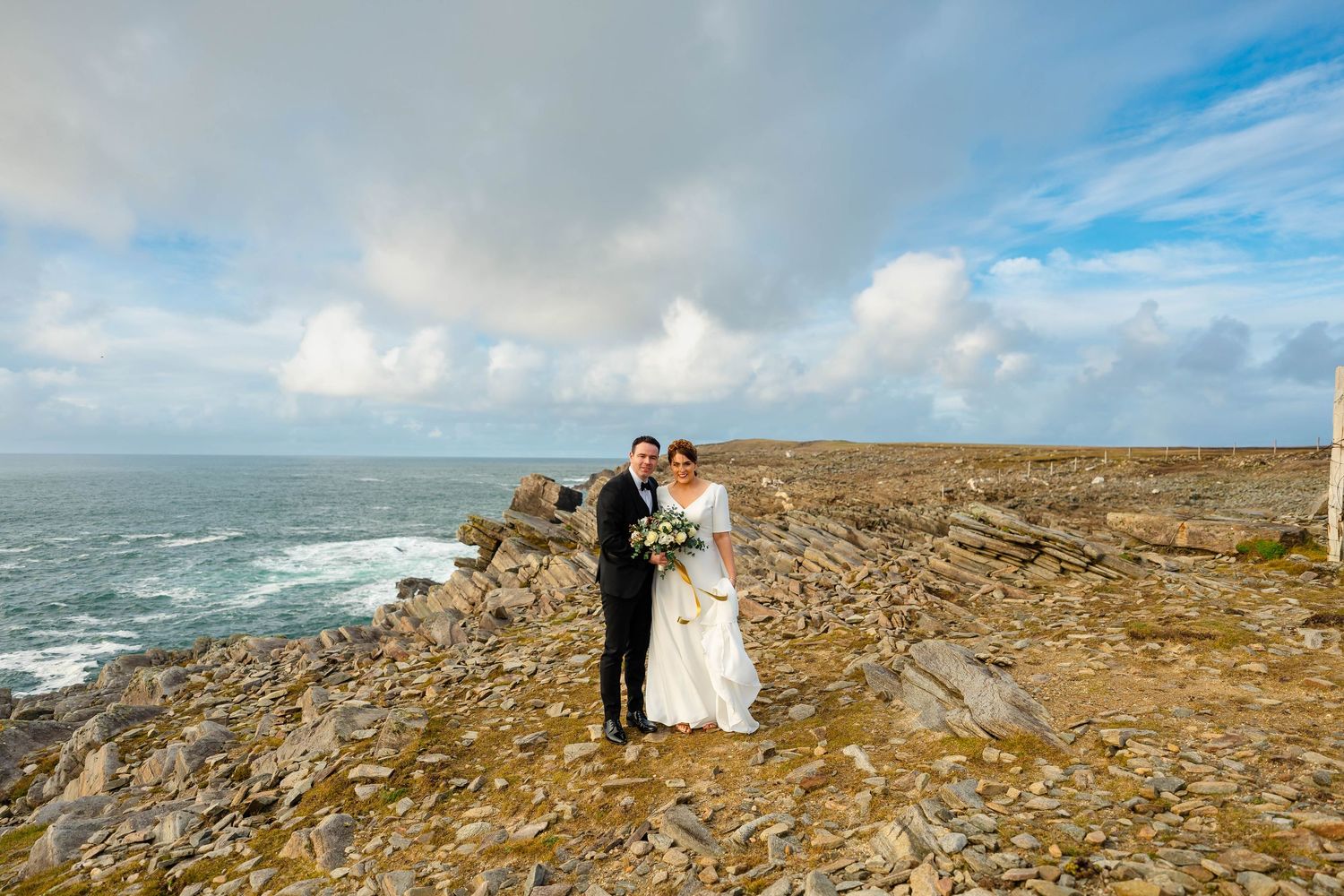 Wedding couple stands on dramatic coastal cliffs overlooking the ocean waves under a blue and cloudy sky.