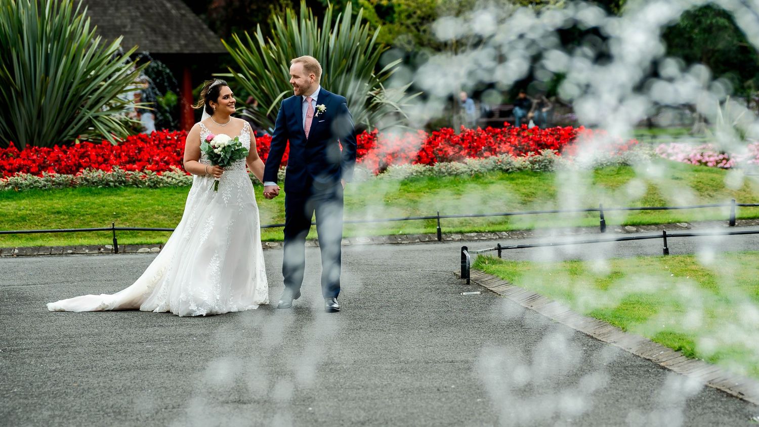A couple walks along a garden path with red flowers and fountain spray in the background.