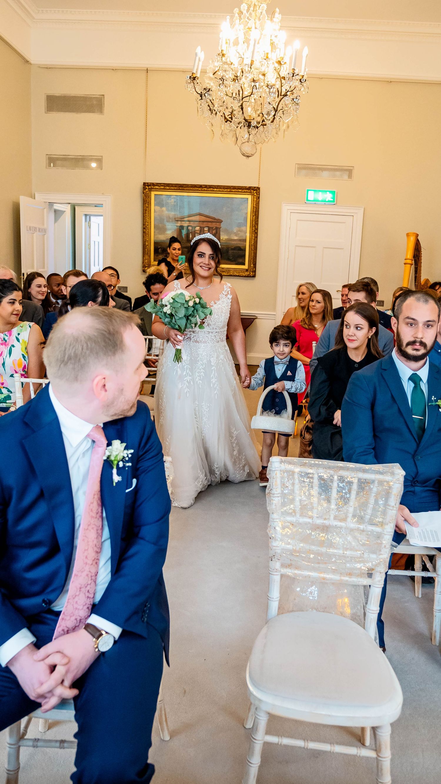 Wedding ceremony setup with white chairs and navy-suited attendees in an elegant cream-colored room.