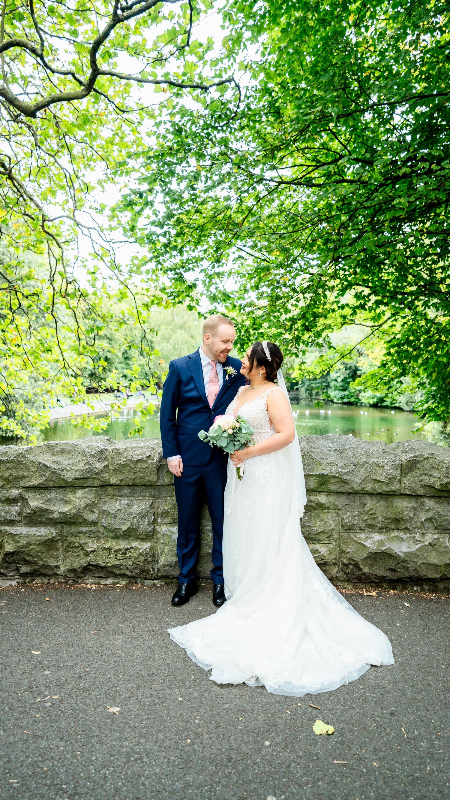 Newlyweds pose together against a stone wall with lush green trees in the background.