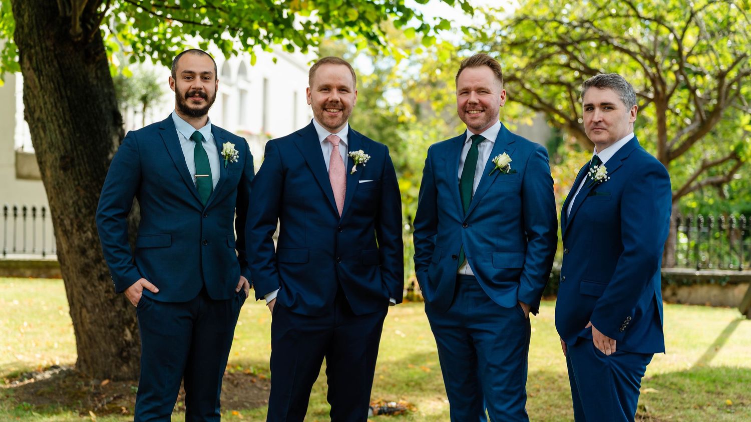 Groomsmen in navy blue suits standing together outdoors under trees.