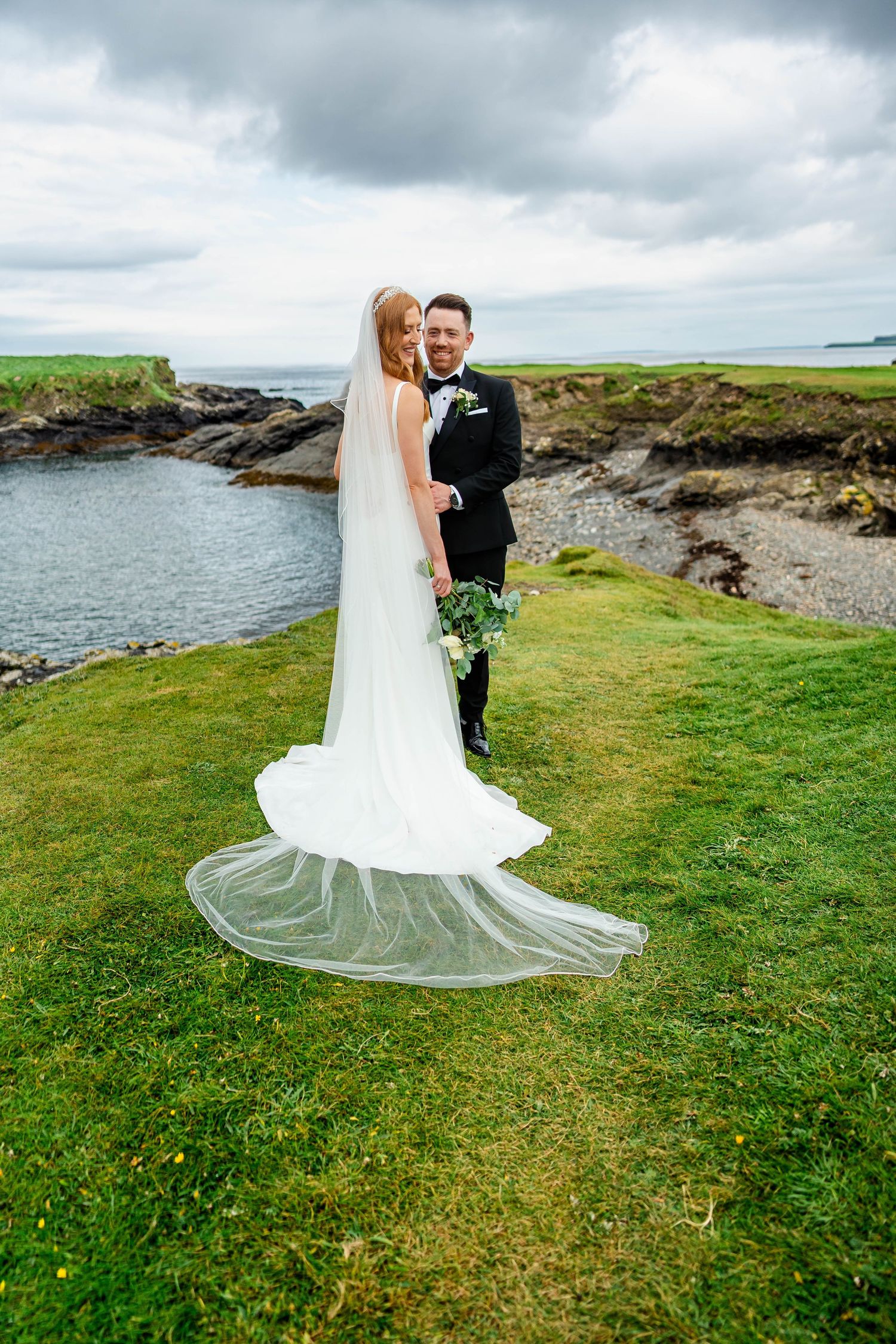 A wedding couple poses on a scenic coastal cliff with green grass and ocean views in the background.