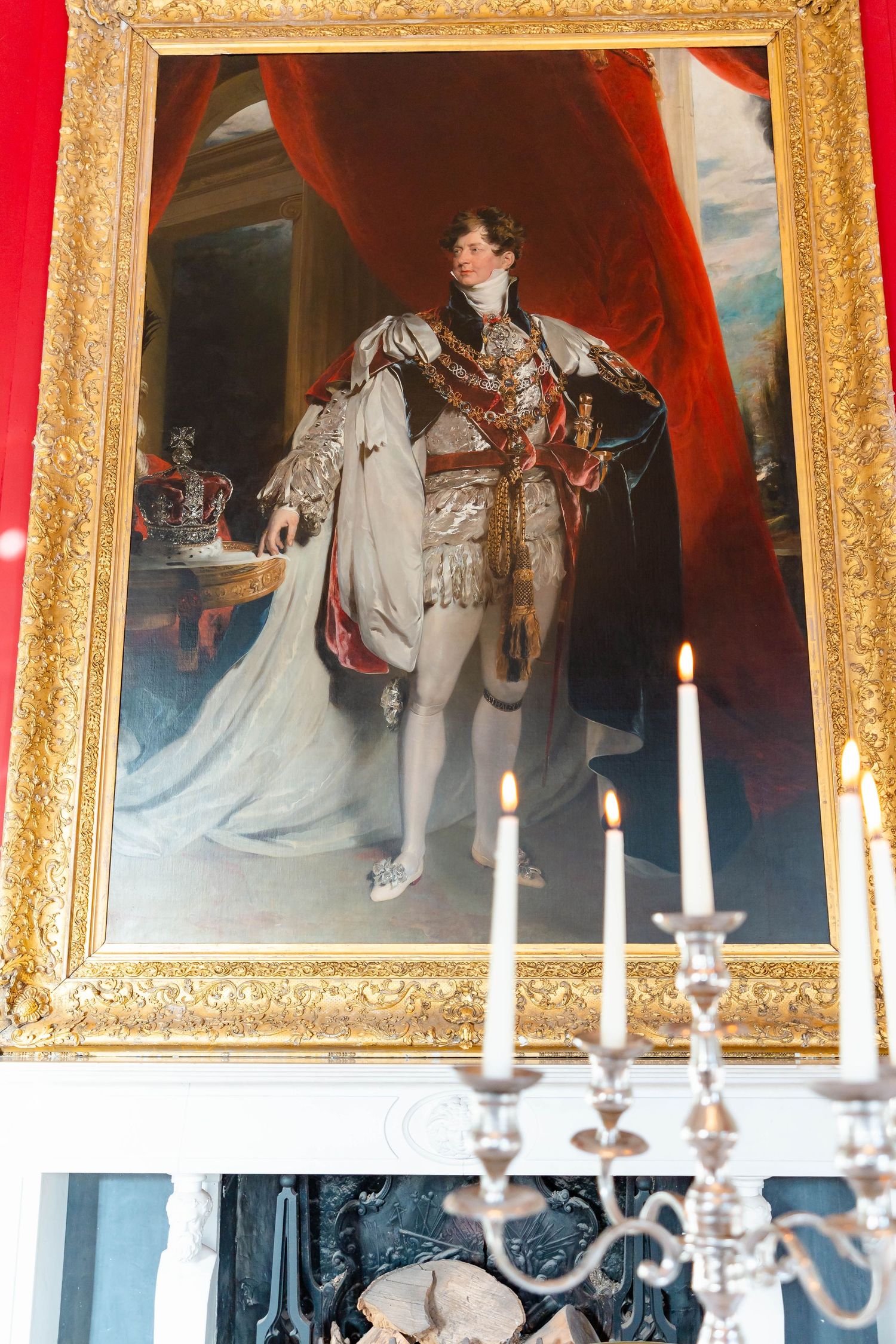 Historic portraits in gilded frames hang on red walls beneath candlesticks in an elegant palace gallery.