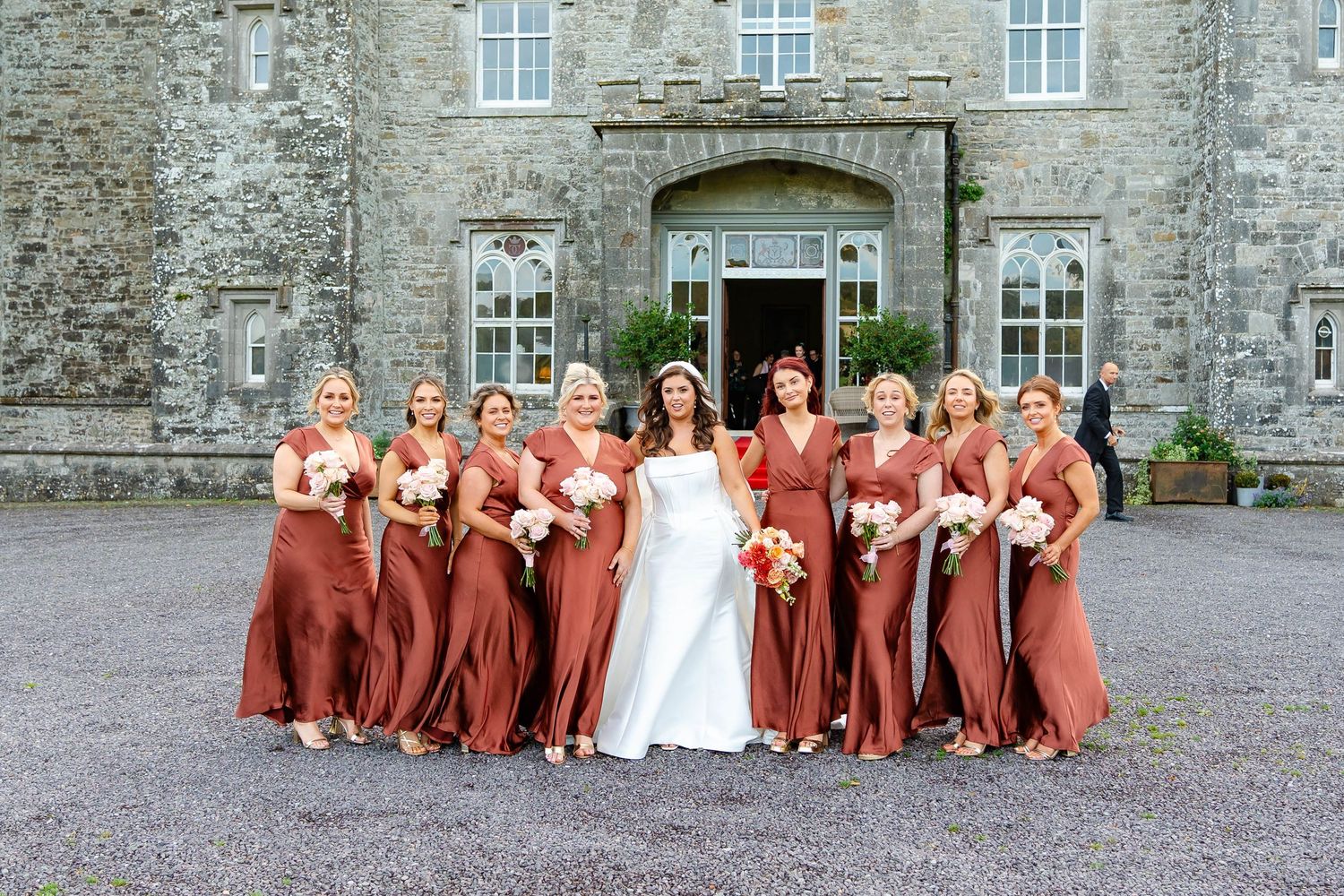 A bride in white stands with her bridesmaids wearing rust-colored dresses outside a historic stone castle.