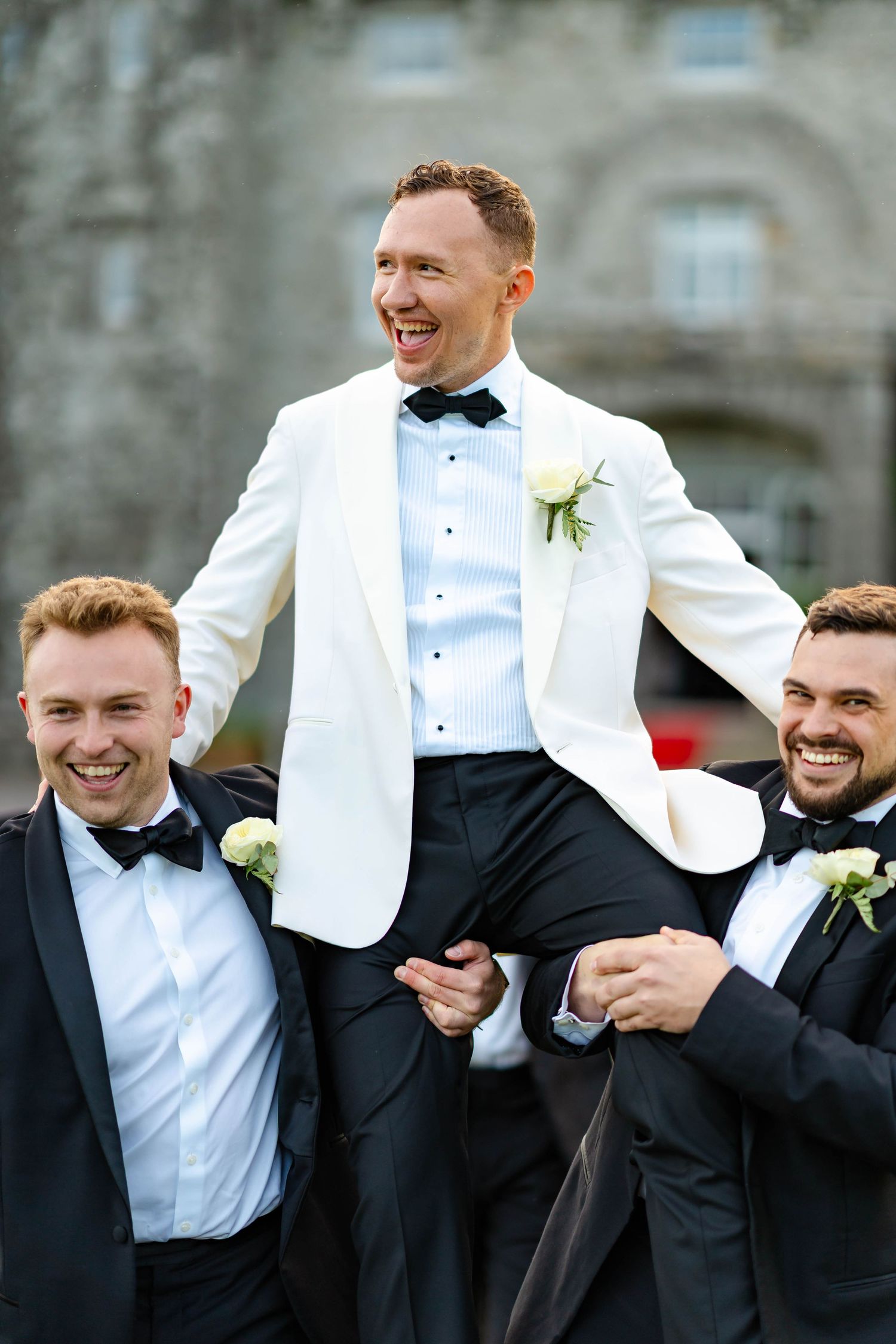 Groomsmen in black and white tuxedos share a candid laughing moment during wedding photos.
