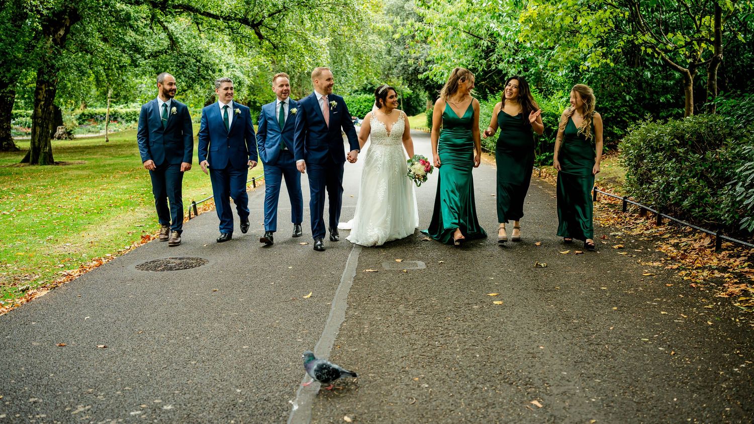 A wedding party walks together along a scenic park pathway in autumn.