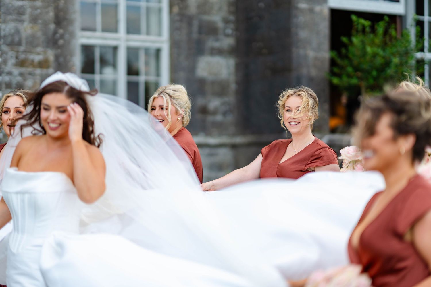 A bride in a flowing white wedding dress shares a joyful moment with bridesmaids in burgundy dresses outdoors.