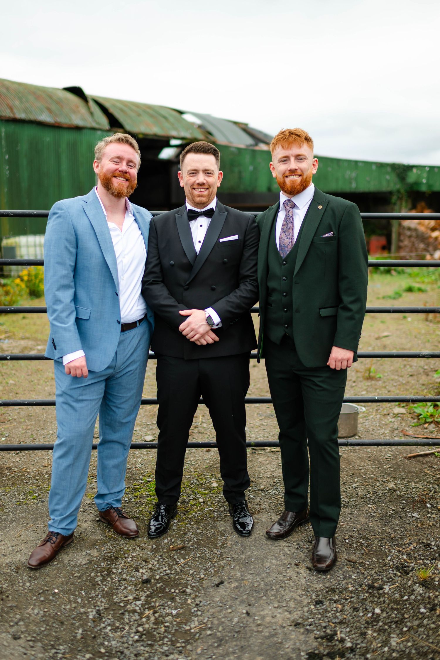 Group of groomsmen in suits posing together on gravel ground outdoors.