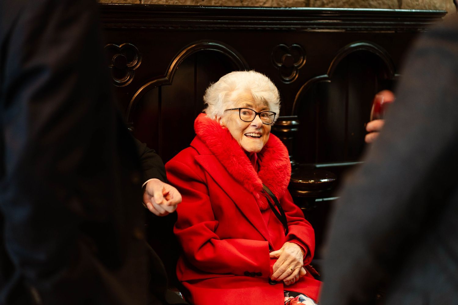 Elderly person wearing bright red coat and glasses smiles while seated indoors.