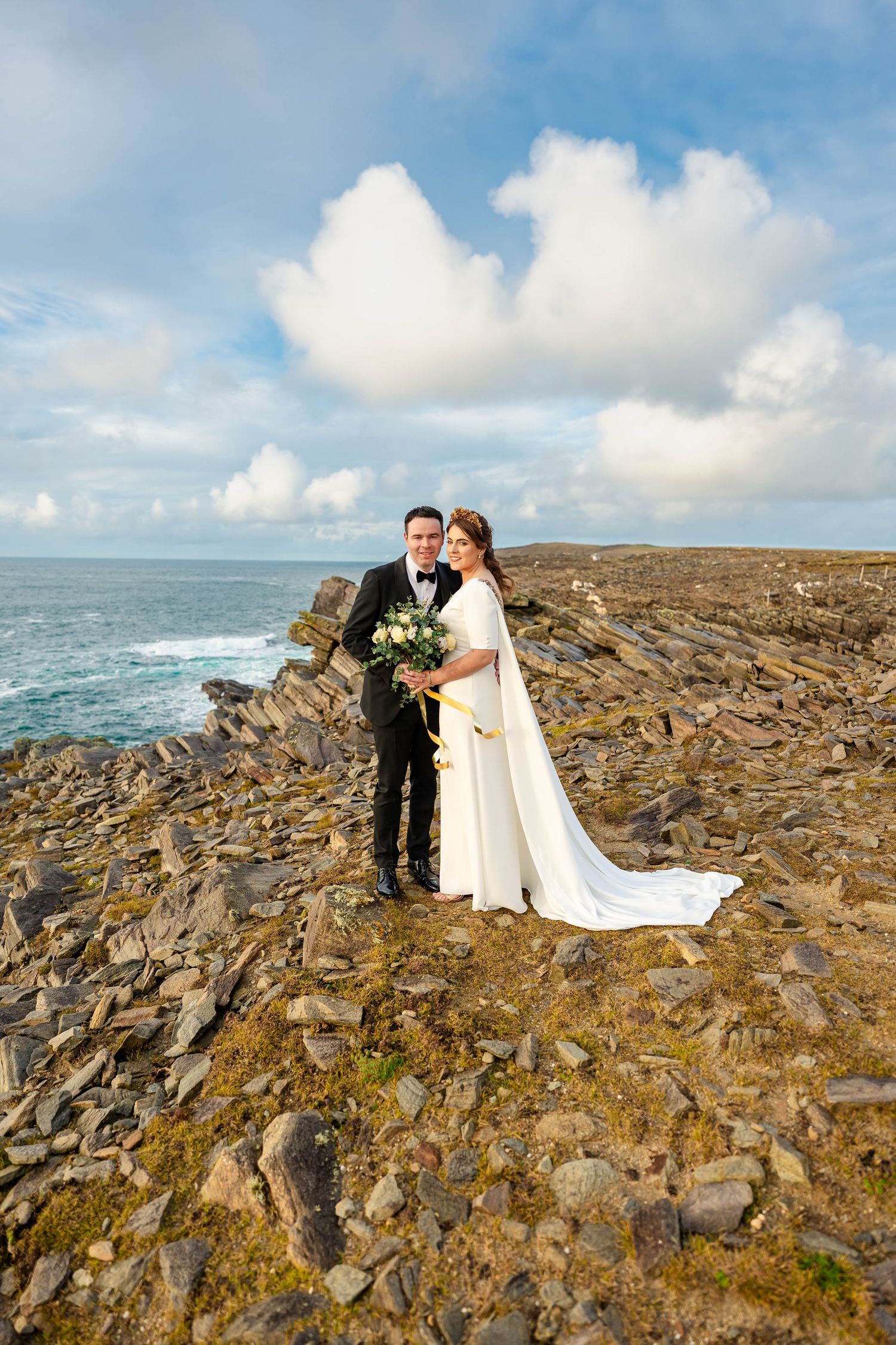 Wedding portrait on rocky coastline with ocean waves and fluffy clouds in background.