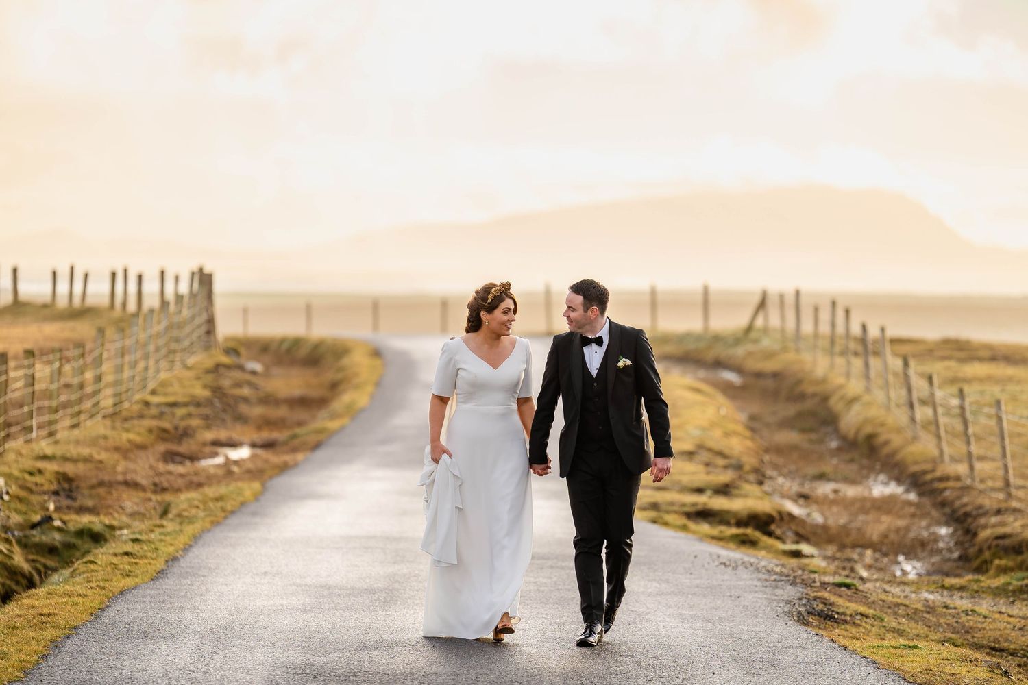 Couple in wedding attire walks along a misty rural road lined with wooden fences.