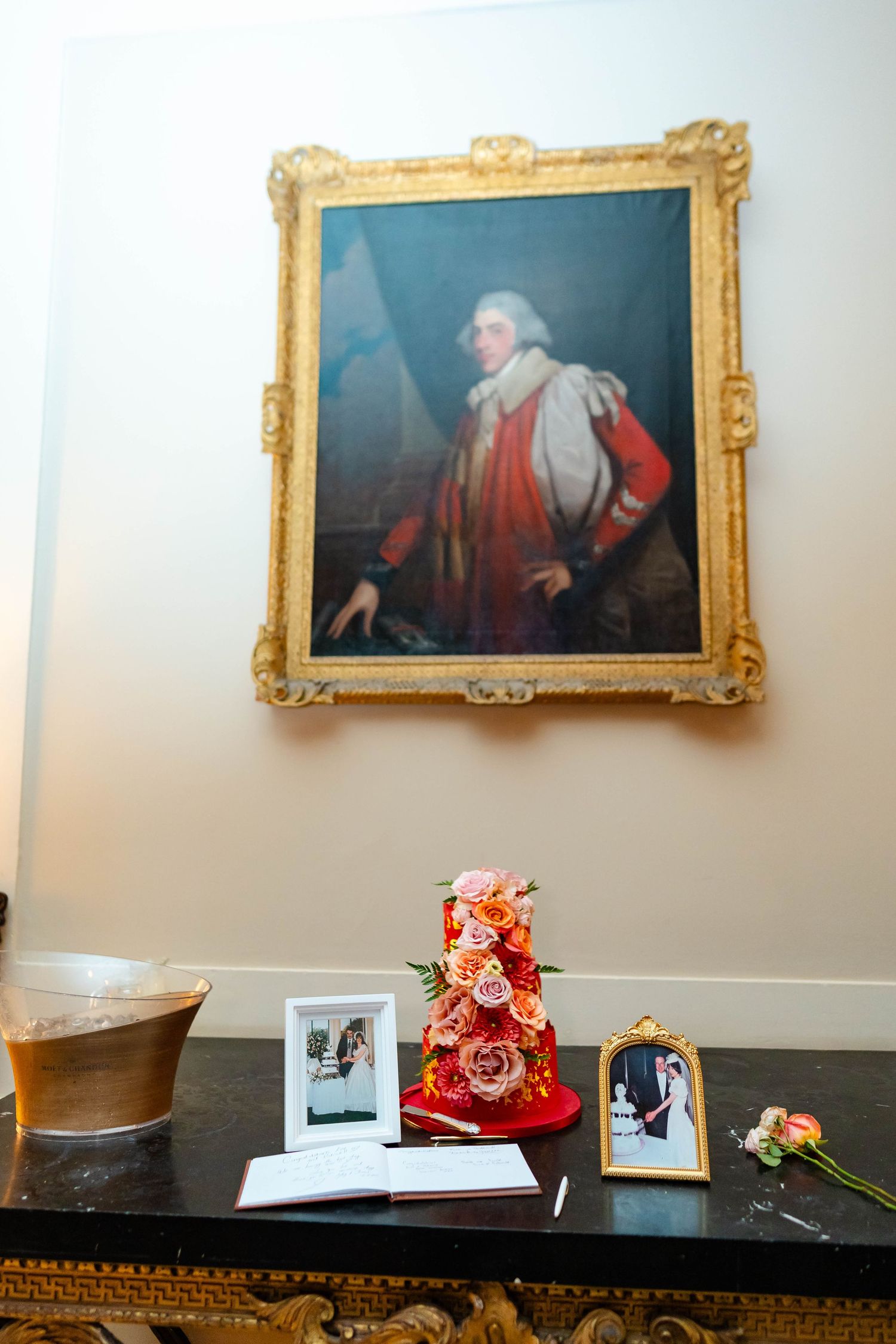 Elegant portrait in ornate gold frame hangs above table with wedding photos and coral-colored floral arrangement.