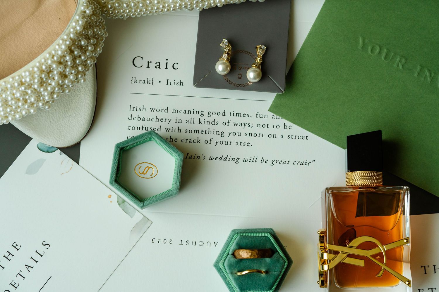 Wedding accessories including pearl necklace, perfume bottles and invitation cards on green background.