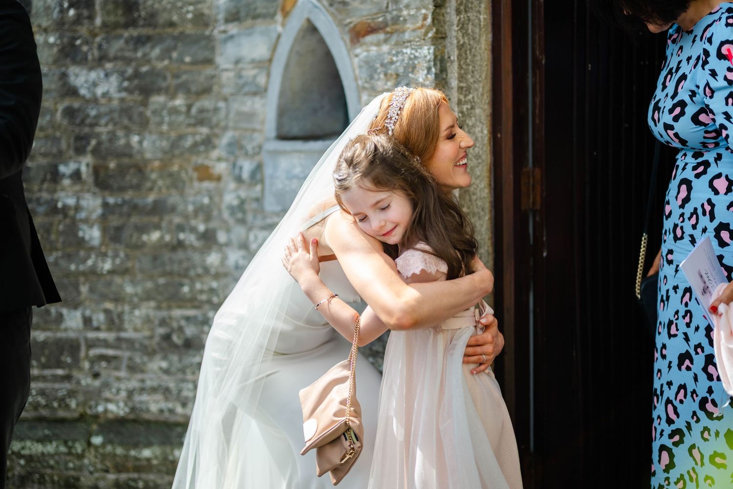 Emotional embrace between wedding attendees outside stone church building.