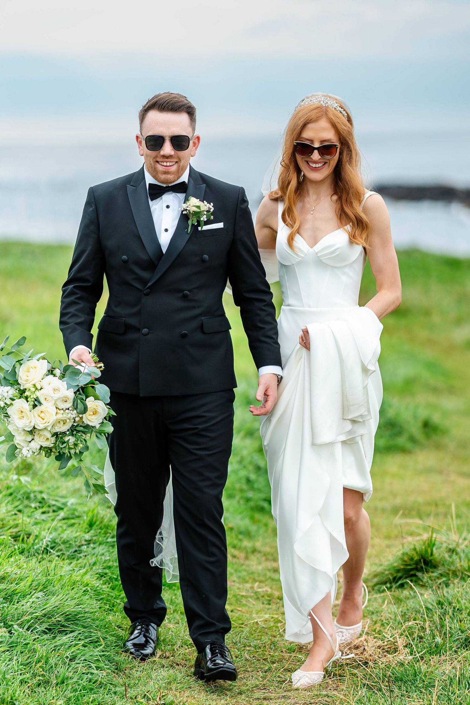 Bride and groom walking along coastal path wearing sunglasses and formal attire.