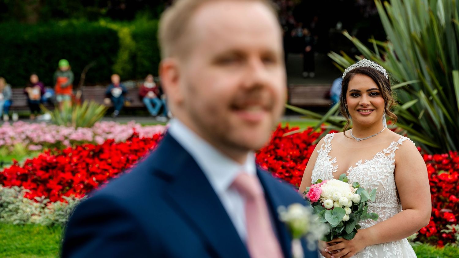 A couple poses in a garden with vibrant red flowers and lush greenery.