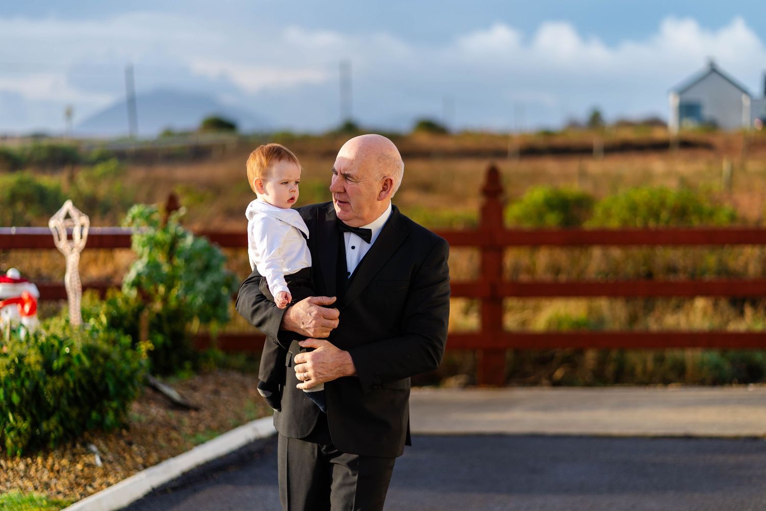Outdoor wedding photos with couple embracing in front of rustic red fence and countryside scenery.