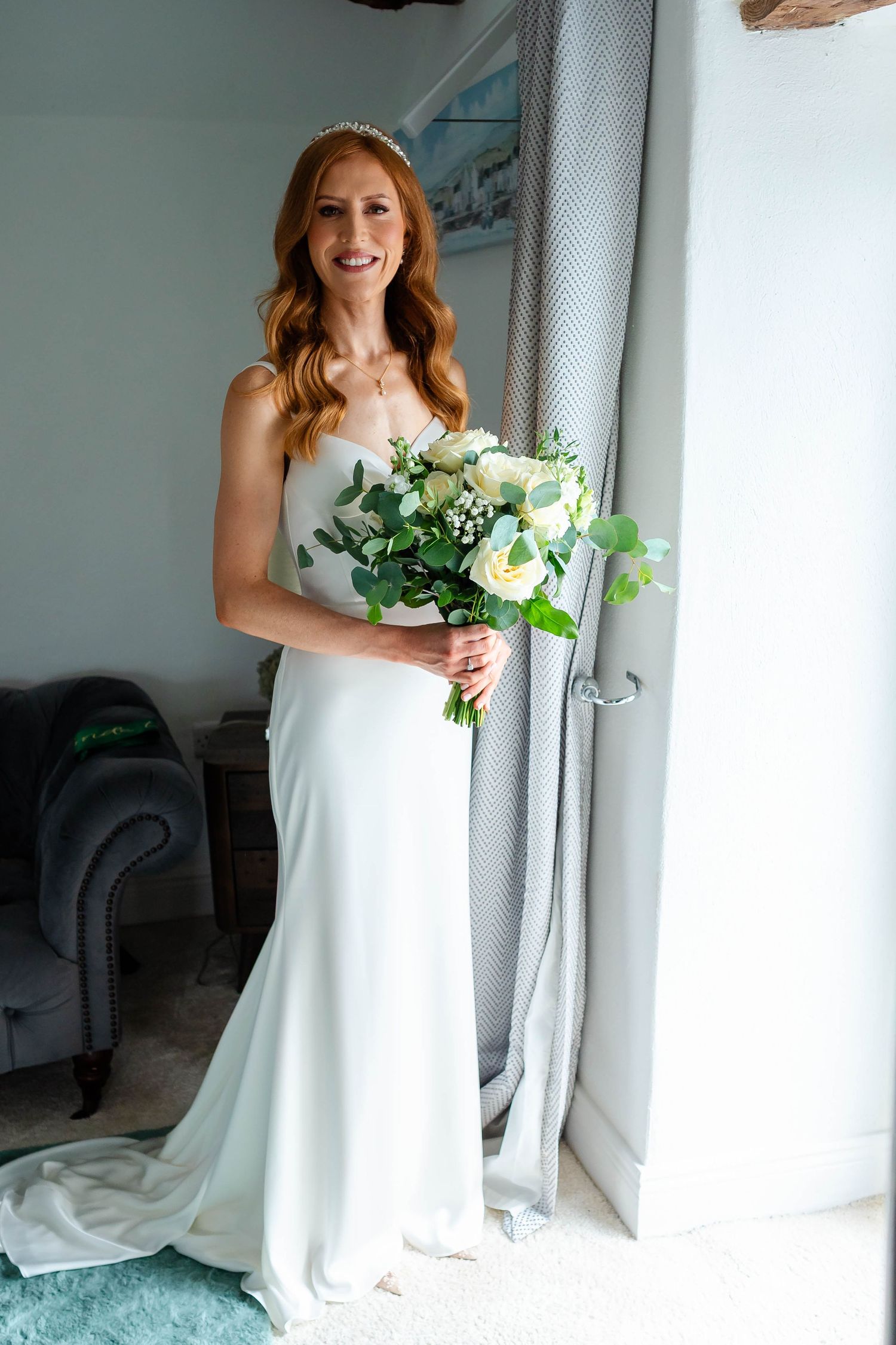 A flowing white wedding gown photographed near a window with a fresh floral bouquet.