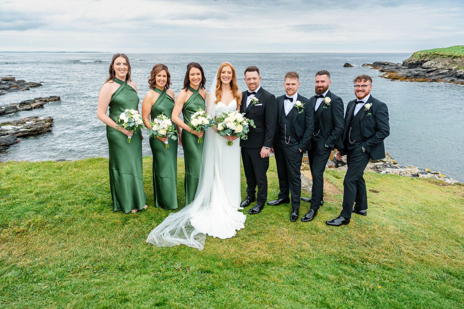 Wedding party in green dresses and black tuxedos poses together on a scenic oceanfront cliff.