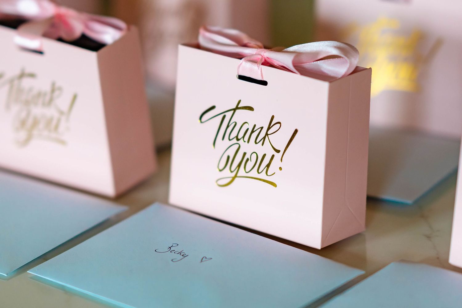 White gift bags with gold lettering arranged in a row on a light surface with soft focus bokeh effect.