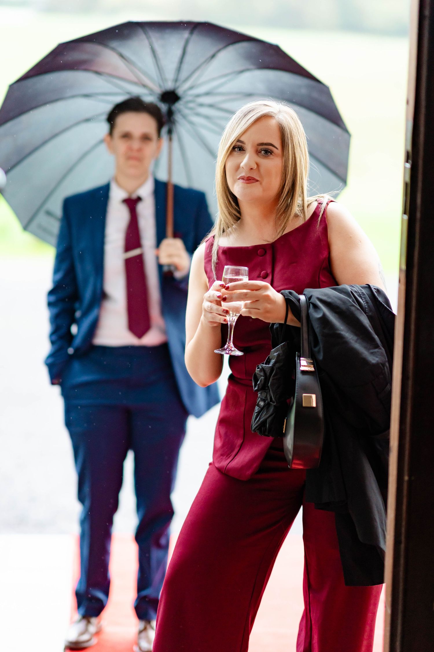 A person in a burgundy dress and another in a navy suit hold umbrellas while enjoying drinks at an outdoor event.