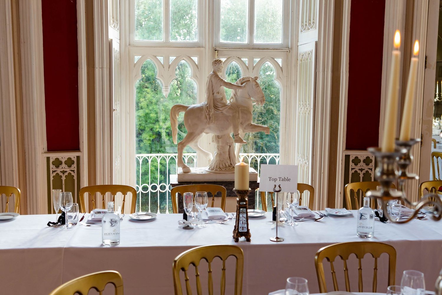 Elegant dining room with white tablecloths, gold chairs, and large windows overlooking a garden setting.