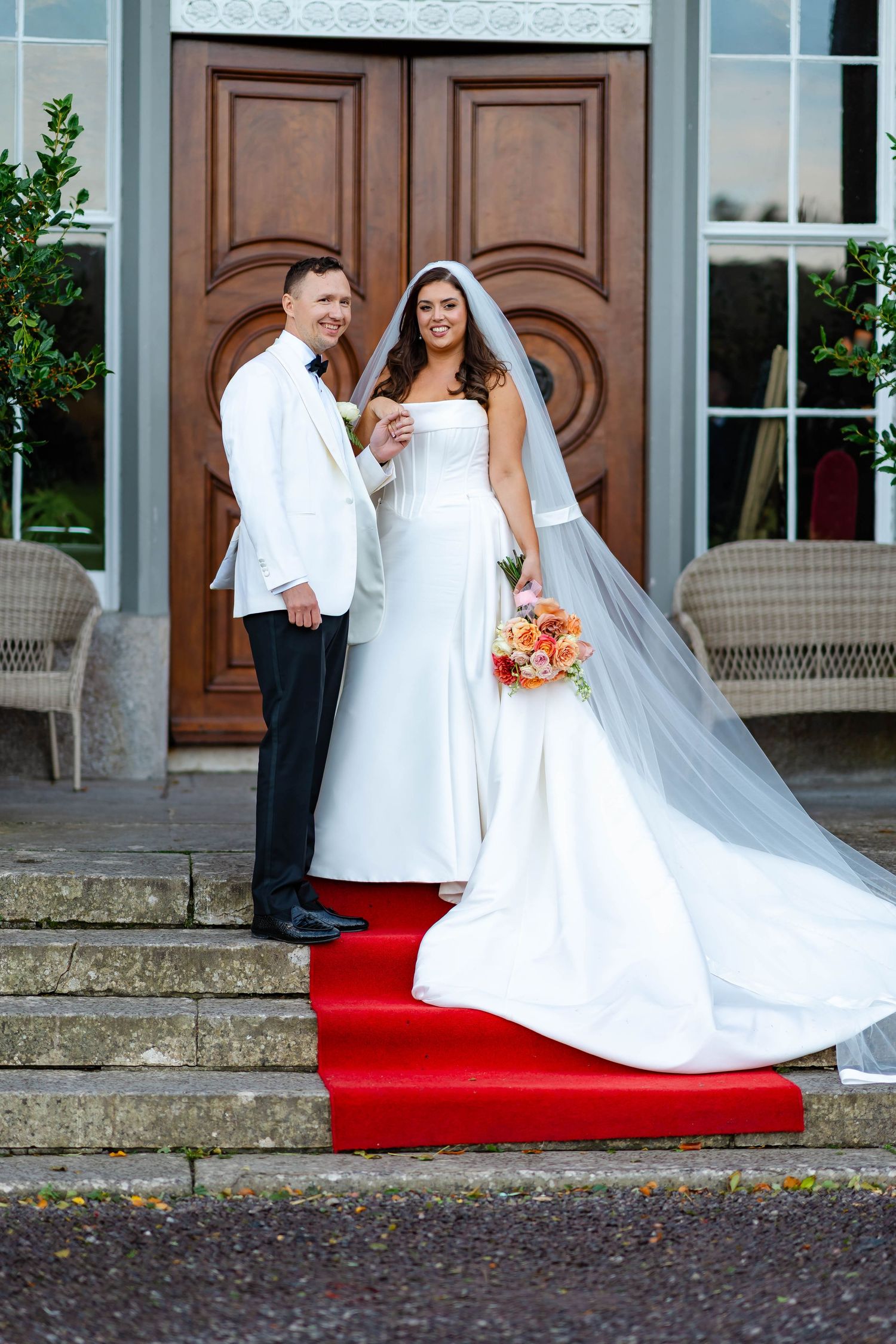 A bride in a flowing white wedding dress with a long train poses on red carpet steps outside elegant wooden doors.