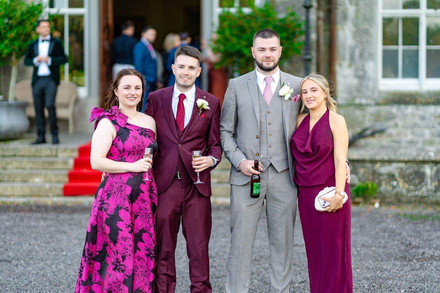 Wedding party members in burgundy and gray attire pose together on gravel path outside stone building.
