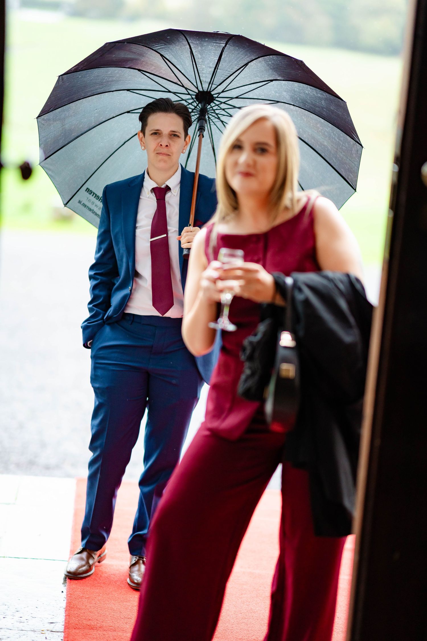 A couple in formal attire holding black umbrellas pose together on a rainy day during an outdoor event.