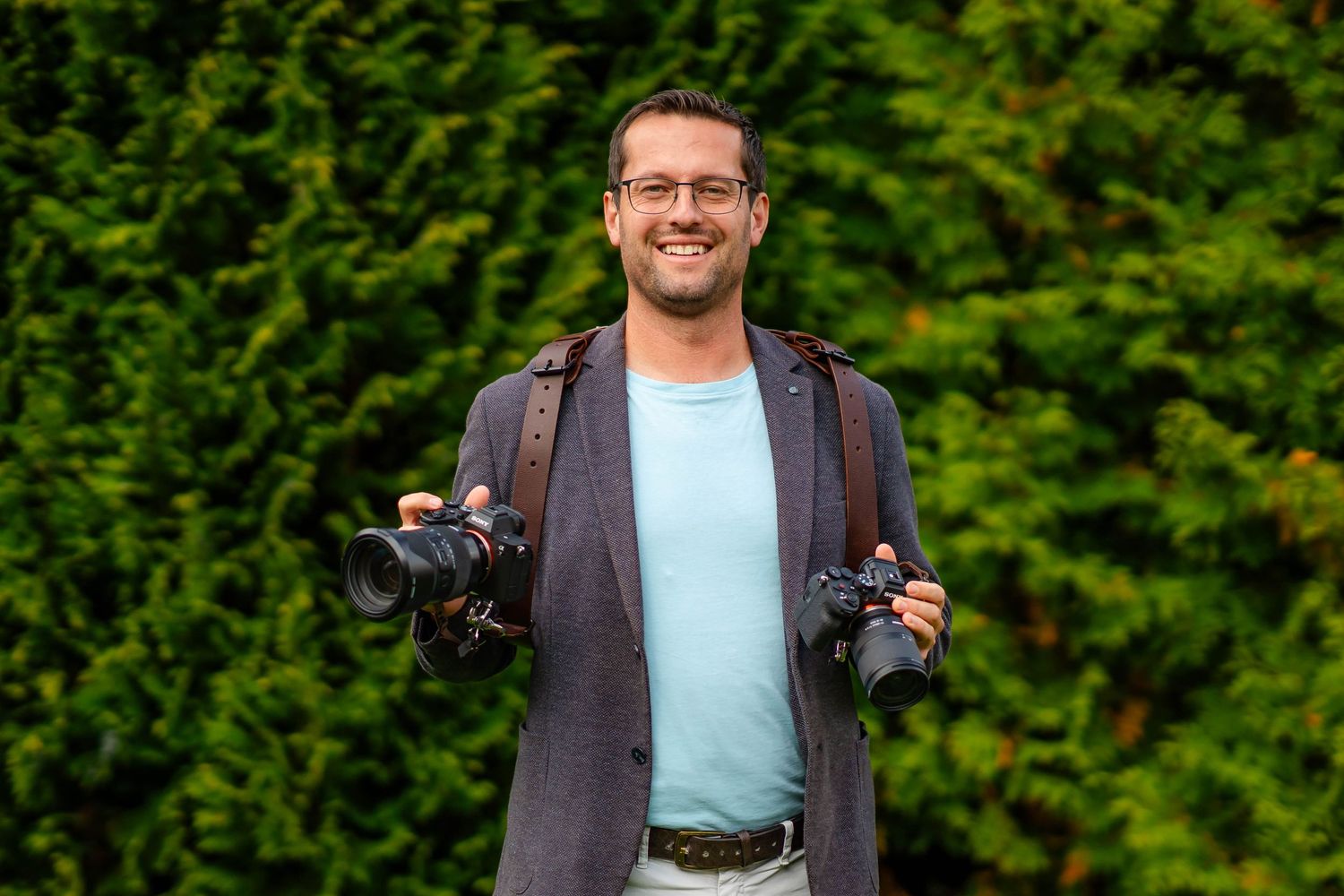 Professional photographer wearing a gray coat holds two cameras against green foliage background.