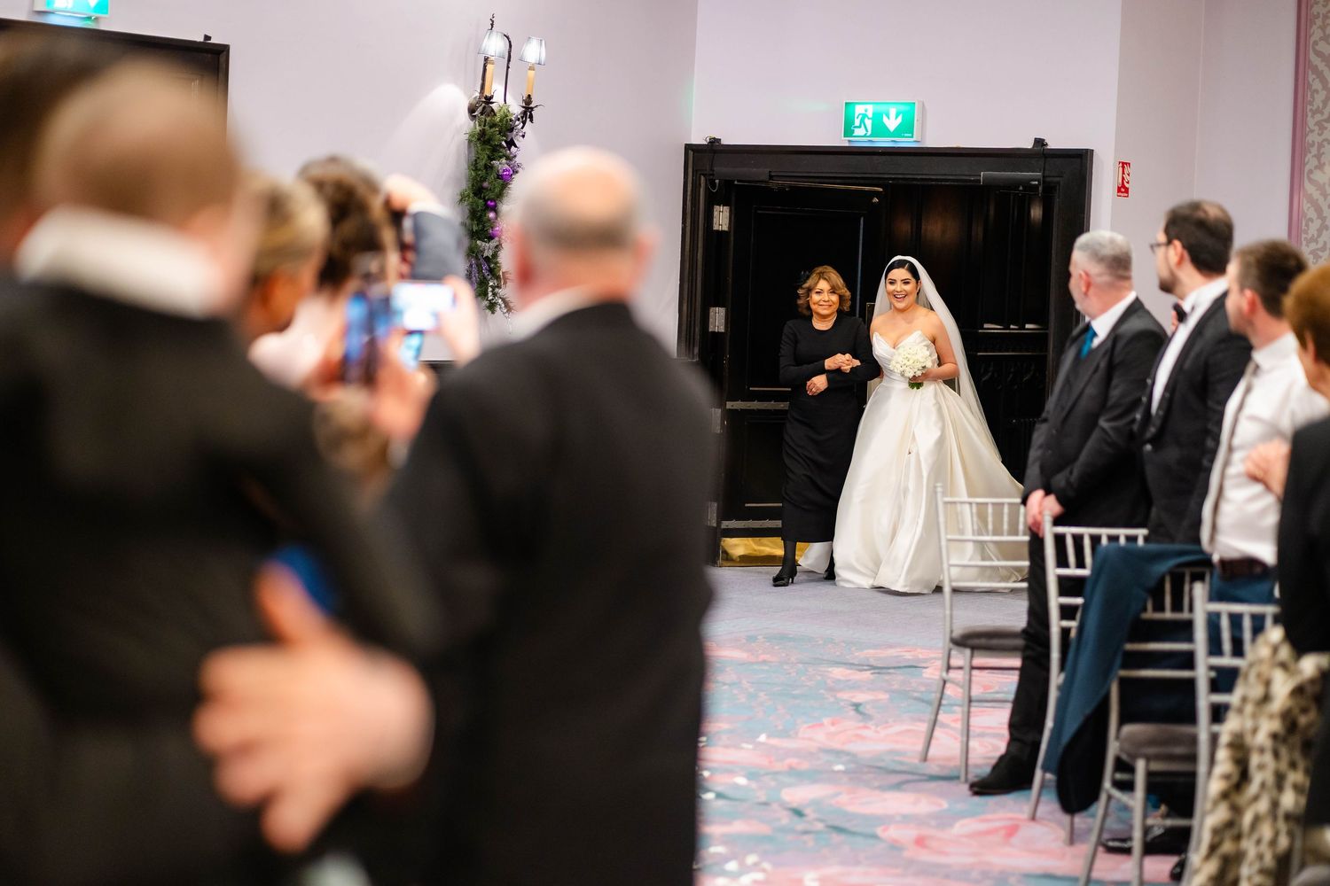 Wedding ceremony entrance captured from behind guests in formal indoor venue.